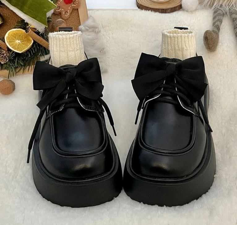Un paio di mocassini in pelle nera liscia su un letto bianco, con un grande fiocco che decora la parte superiore e piccoli calzini beige che riempiono la scarpa.