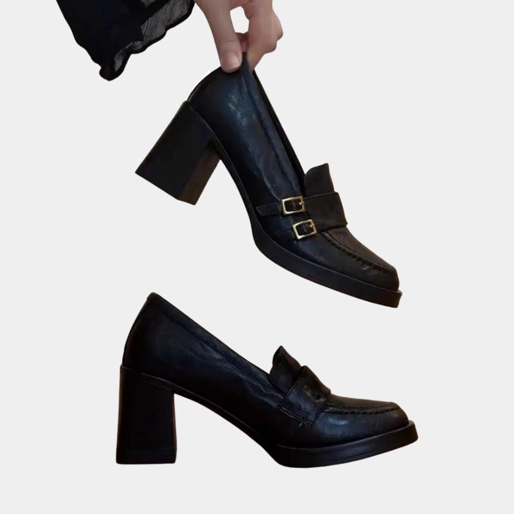 Paio di mocassini con tacco alto neri, di cui una scarpa appoggiata di profilo e la seconda tenuta tra le dita di una donna