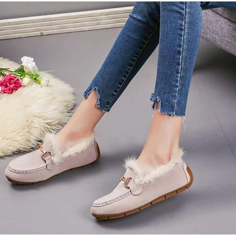 Un paio di mocassini invernali in pelle scamosciata beige con pelliccia visibile, indossati con jeans e lo stesso modello in nero accanto. Il pavimento è grigio e presenta un pezzo di tappeto di pelliccia beige.