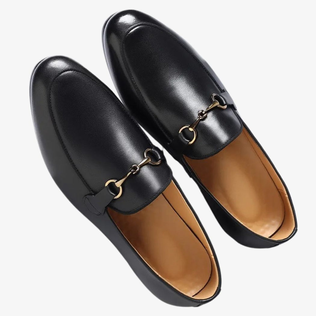 Mocassino slip-on nero con fibbia dorata in pelle vegana su sfondo bianco sporco.