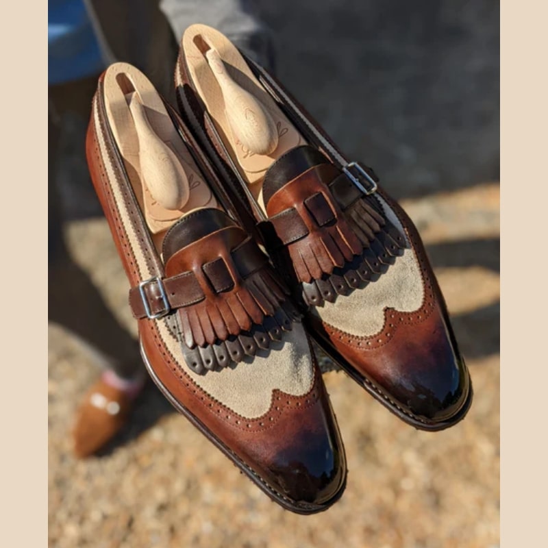 Un paio di mocassini semi-lucidi in pelle liscia e camoscio sintetico marrone e beige, con frange bicolore e chiusura con cinturino e fibbia nella parte superiore.