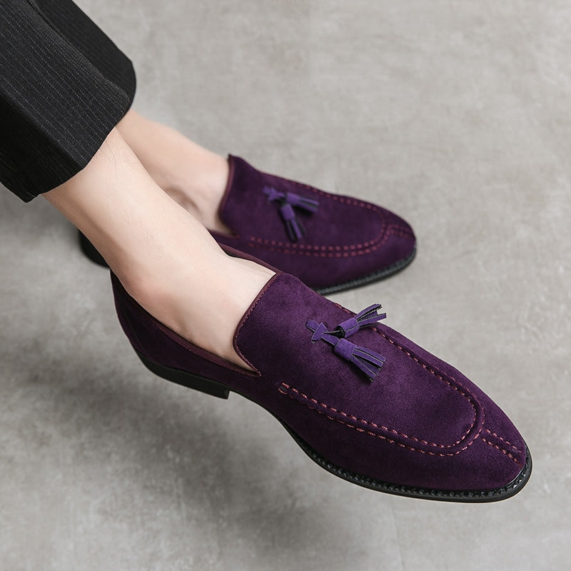 Mocassini in pelle scamosciata viola con nappe decorative ai piedi di un uomo a gambe incrociate che indossa pantaloni da abito neri con sottili strisce bianche.