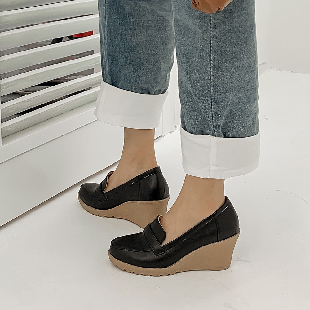 Foto di un paio di mocassini con zeppa neri indossati da una donna in jeans su un pavimento grigio