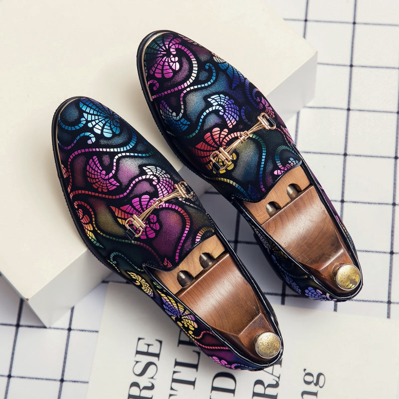 Un paio di mocassini da sera con arabeschi multicolori e punte d'argento è esposto su una superficie a scacchi con scritte in bianco e nero. Ci sono alberi di scarpe in legno con un bottone d'oro all'interno delle scarpe.