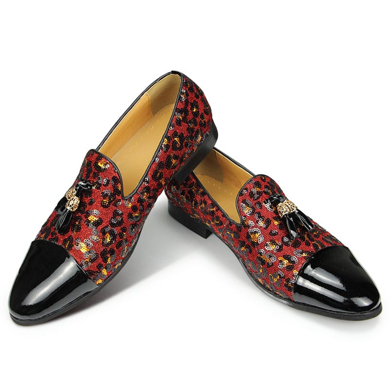 Su uno sfondo bianco, un paio di mocassini con una parte anteriore in vernice nera, il resto della scarpa in paillettes con stampa leopardo in rosso e nappe sulla parte superiore. L'interno è beige