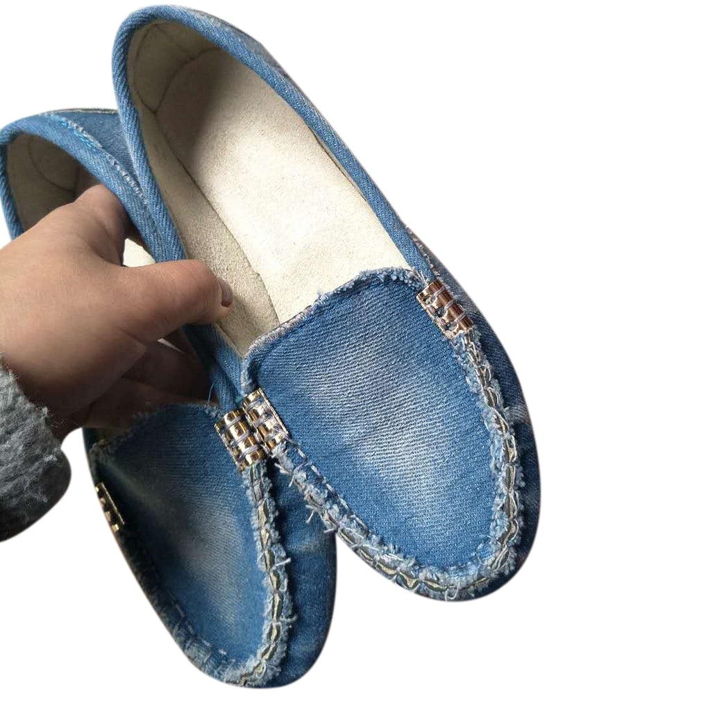 Un paio di mocassini morbidi in denim blu sbiadito con suole in gomma beige con borchie, indossati a piedi nudi da una donna che indossa jeans strappati blu con caviglie esposte