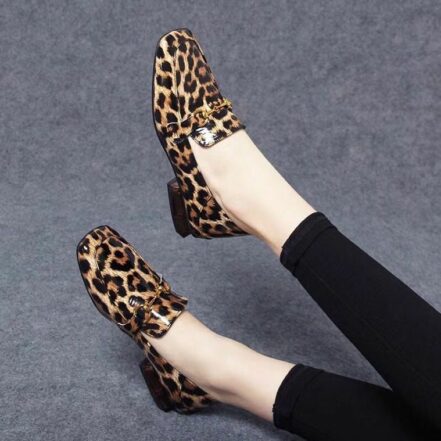 Su uno sfondo grigio, un paio di gambe con leggings neri e mocassini lucidi con tacchi quadrati leopardati