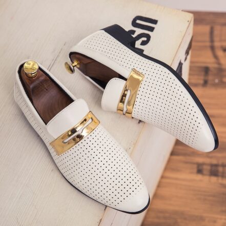 Su una scatola di legno chiaro, un paio di mocassini bianchi allungati con piccoli fori e un cinturino dorato sulla parte superiore. All'interno delle scarpe ci sono degli alberi da scarpe in legno con un bottone d'oro sul tallone.