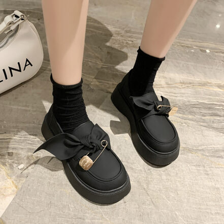 Mocassini rotondi, lisci, dalla suola spessa, con un grande fiocco e una finta spilla che decora la parte superiore della scarpa, ai piedi di una donna in piedi su un pavimento di marmo bianco e che indossa piccoli calzini neri.