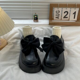 Un paio di mocassini in pelle nera liscia su un letto bianco, con un grande fiocco che decora la parte superiore e piccoli calzini beige che riempiono la scarpa.