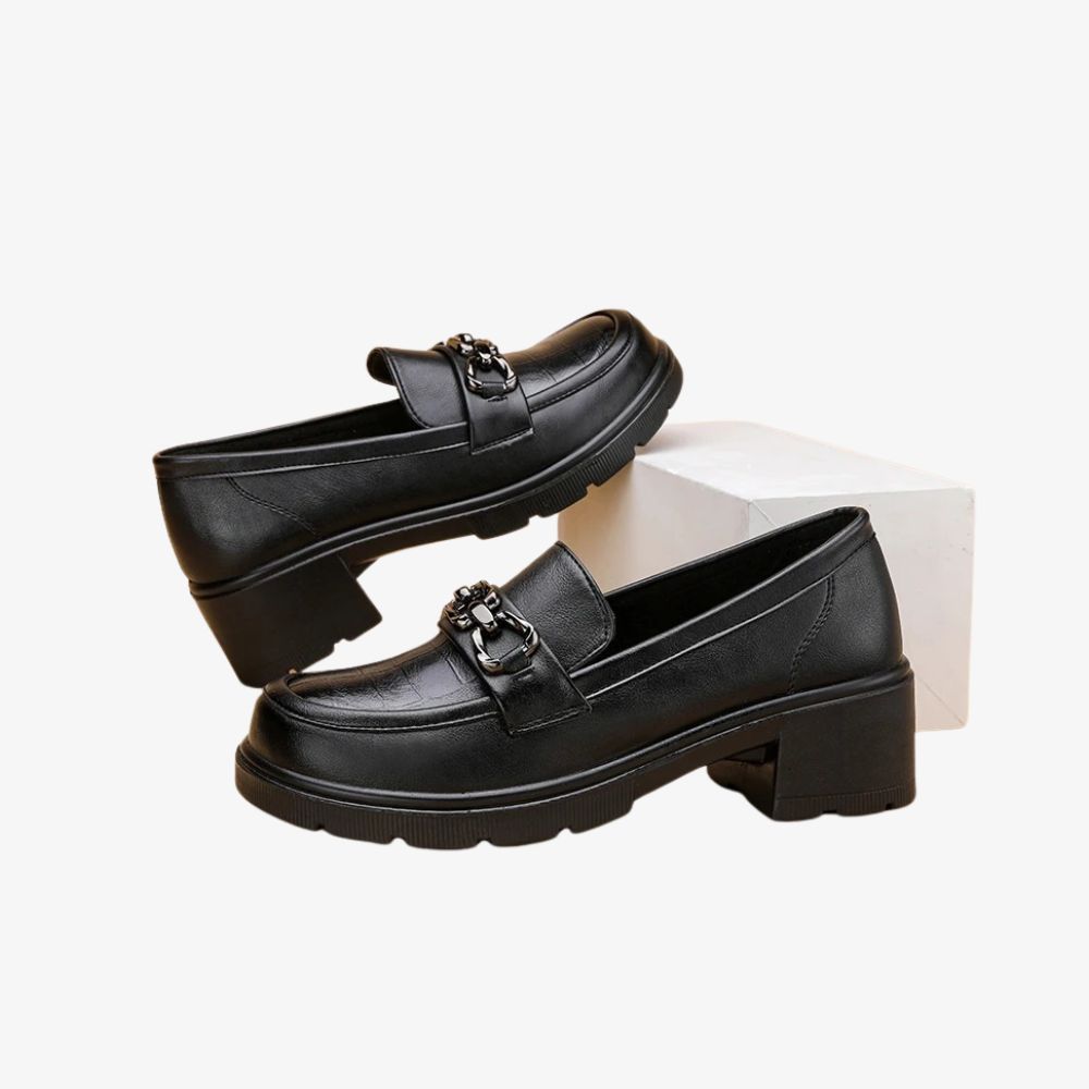 Paire de mocassins noirs à talons avec une chaussure posée sur un bloc blanc.