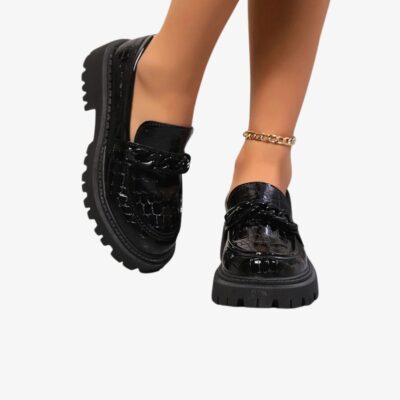 Paire de mocassins noirs vernis aspect croco, portés par une femme dont on ne voit sur les chevilles. Elle porte un bracelet de cheville.
